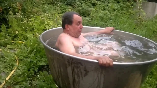 Naked old man rolls around in outside bath tub.